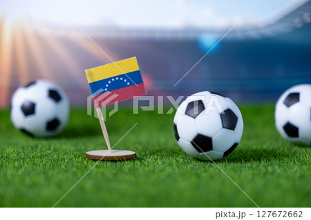 Small Venezuelan national flag stands prominently on a green football pitch, surrounded by miniature soccer balls, symbolizing national pride and international athletic events Small Venezuelan national flag stands prominently on a green football pitch, surrounded by miniature soccer balls, symbolizing national pride and international athletic events 127672662