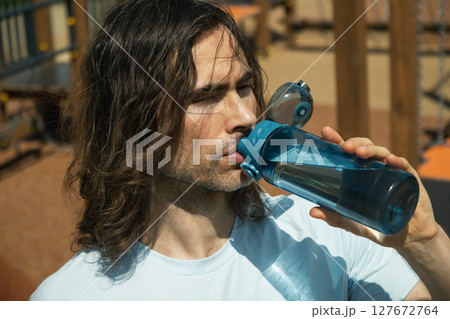 Fit brunette caucasian young man drinking water in blue t-shirt on outdoor sports ground after outdoors work out. Water balance for heat season. Close up. Fit brunette caucasian young man drinking water in blue t-shirt on outdoor sports ground after outdoors work out. Water balance for heat season. Close up. 127672764