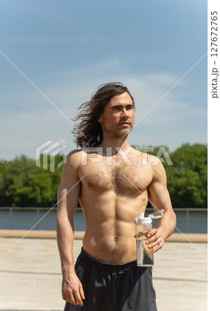Shirtless fit brunette man with curly long hair holding a water bottle while standing outdoors on a sunny day against clear blue sky and lush green trees as nature background. Heat summer. 127672765