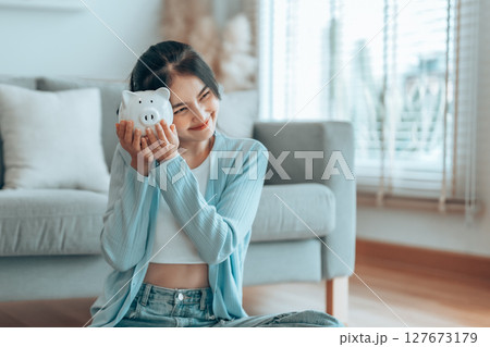 Young Asian woman holding piggy bank and sitting on the floor at home 127673179