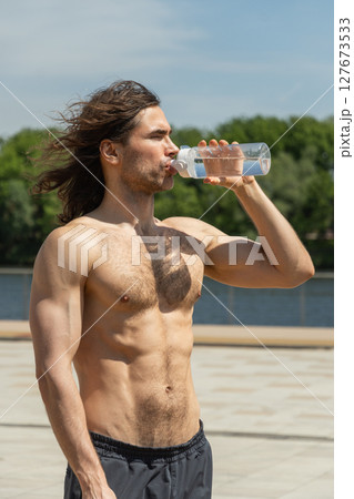 Shirtless fit brunette man drinking water in sunny day against clear blue sky. Heat summer. 127673533