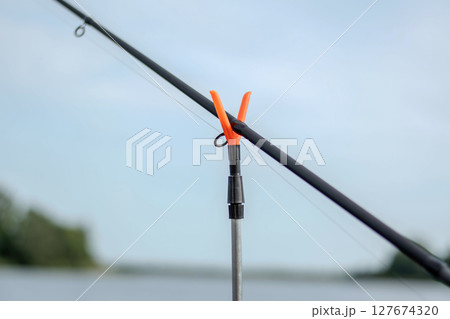 Close-up of a fishing rod secured by a neon orange holder, set against a backdrop of water and green vegetation. 127674320