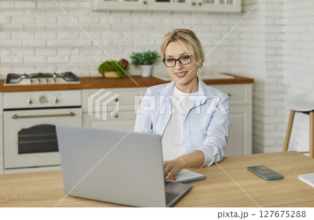 Woman working on a laptop sitting at the desk on kitchen and looking at computer monitor screen 127675288