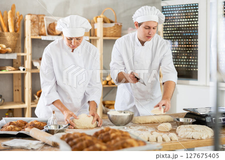 Focused female and guy baker in white uniform working at bakery, kneading and cutting dough into pieces Focused female and guy baker in white uniform working at bakery, kneading and cutting dough into pieces 127675405