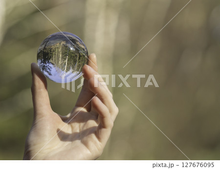 Hand holding glass ball with tree reflection in green forest. Transparent crystal sphere symbolizing 127676095