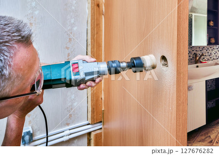 Completing hole cutting process man holds power drill with wood core stuck inside hole saw after creating opening for latch on wooden door surface. Completing hole cutting process man holds power drill with wood core stuck inside hole saw after creating opening for latch on wooden door surface. 127676282