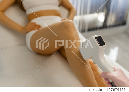 A female cosmetologist does laser hair removal on the slender legs of a beautiful young woman lying on a medical couch in a beauty salon. Cosmetology, hair removal and spa concept. Body care. Close-up 127676351