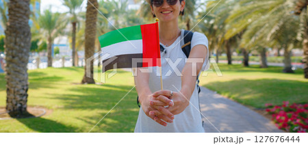 woman holding tiny flag of UAE against Abu Dhabi skyline, Promenade in Abu Dhabi. United Arab Emirates, Middle East. Immigration, student education and Arab emirates citizenship concept 127676444