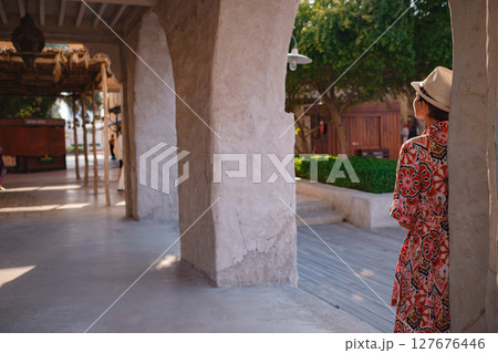 Young woman in an ethnic dress and hat exploring Al Seef Park in Dubai. Surrounded by traditional Emirati architecture and modern vibes, she enjoys the charm of Dubai Creek's cultural heritage. 127676446