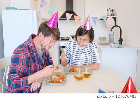 Young couple celebrates cozy birthday at home, enjoying cake and laughter in warm setting. Family are eating pasta and drinking wine to celebrate, enjoy meal and cheers Young couple celebrates cozy birthday at home, enjoying cake and laughter in warm setting. Family are eating pasta and drinking wine to celebrate, enjoy meal and cheers 127676483