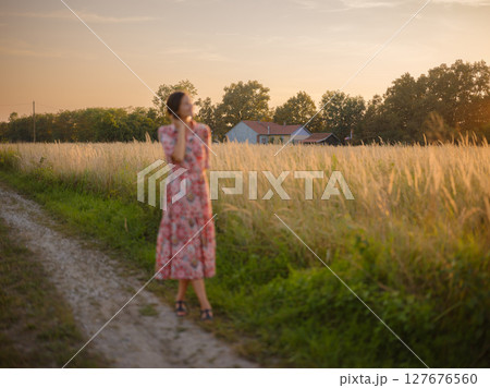 Young woman walking through picturesque European field in late summer. Golden sunlight, lush greenery, and serene rural atmosphere create peaceful countryside scene. 127676560