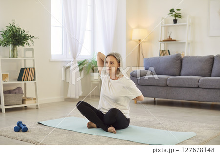 Focused mature elderly woman sitting on yoga mat and warming up with stretching exercises at home. Focused mature elderly woman sitting on yoga mat and warming up with stretching exercises at home. 127678148