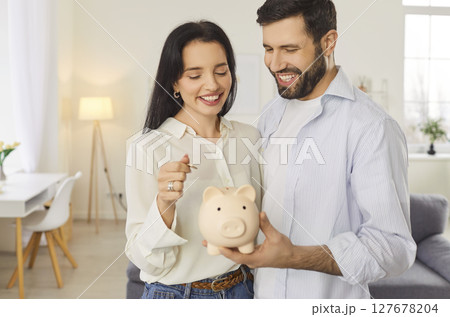 Happy young smiling couple holding piggy bank and putting money coin standing at home together. Happy young smiling couple holding piggy bank and putting money coin standing at home together. 127678204