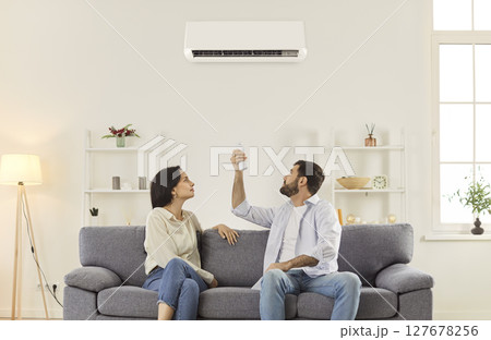 Young couple sitting on sofa in living room, turning on air conditioner with remote controller. 127678256
