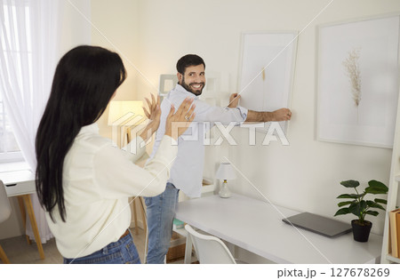 Happy young couple decorating new home, hanging picture on wall with wife guiding smiling husband. 127678269