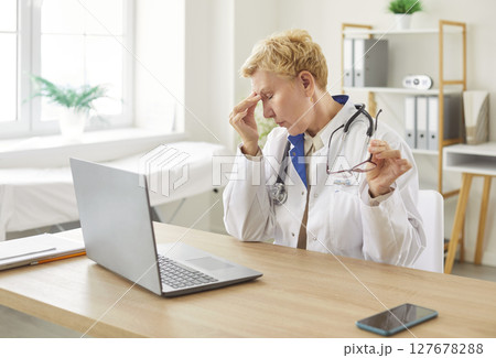 Tired female doctor resting at desk, putting off glasses and rubbing nose after working on laptop. 127678288