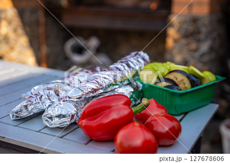 Foil-wrapped fish and fresh vegetables arranged on a table before grilling in the backyard. Outdoor cooking, natural food preparation, and homemade barbecue setup. 127678606