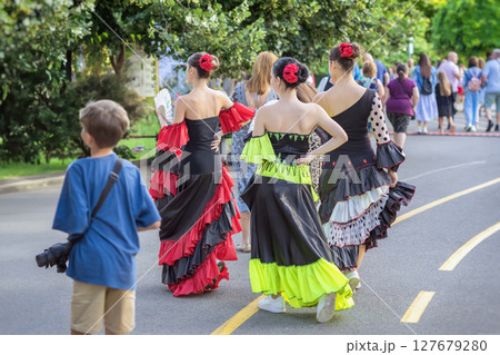 Folk ethnic flamenco dancers walking along street with people. Group of flamenco women dancers in bright dresses walking among crowd at cultural festival in park setting. Spanish Culture Festival 127679280