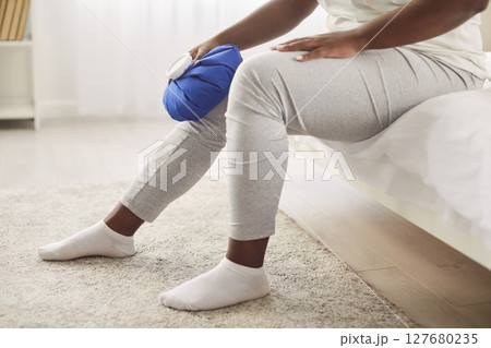 Cropped portrait of african american woman applying cold compress to leg sitting on bed at home Cropped portrait of african american woman applying cold compress to leg sitting on bed at home 127680235