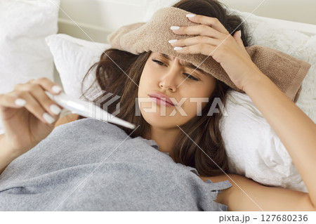 Sick young woman lying in bed under plaid, suffering from cold or flu and checking her temperature. 127680236