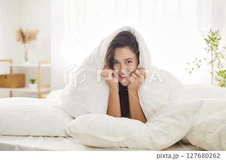 Young woman smiling while lying on soft pillow in her cozy bed with warm blanket covering her head. 127680262