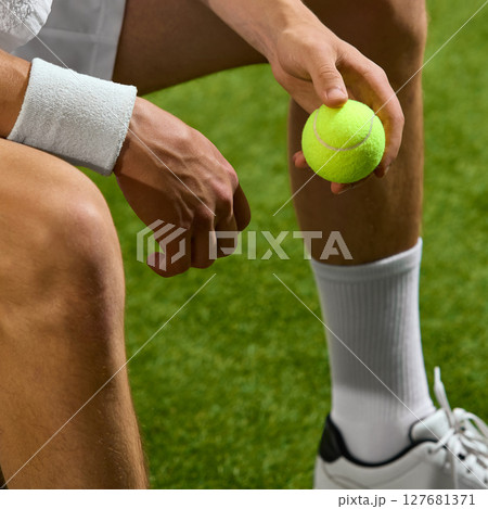 Tennis player preparing for match holding ball seated on grass court with wristband and sports shoes 127681371