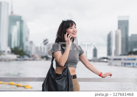 Happy young Asian woman talking on smartphone with cheerful expression at an urban city waterfront. Modern lifestyle and travel tourism concept in metropolis with buildings Happy young Asian woman talking on smartphone with cheerful expression at an urban city waterfront. Modern lifestyle and travel tourism concept in metropolis with buildings 127681468