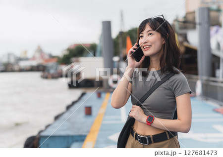 Happy solo Southeast Asian woman, teenager blogger, on world travel tourism experience in Thailand. She wait for boat on Chao Phraya River in Bangkok using smartphone to express joy to friend 127681473