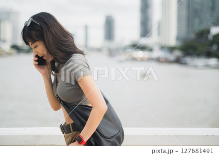 Solo southeast asian woman, teenager world travel explorer, on tourism trip in Asia. blogger has serious phone call with friend via smartphone by Chao Phraya River in Bangkok, Thailand 127681491