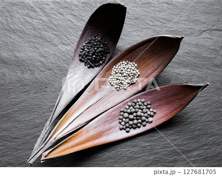 Black, White and Fragrant Piles of Pepper Grains in Dry Leaf Funnels on Black Stone Background Surface 127681705