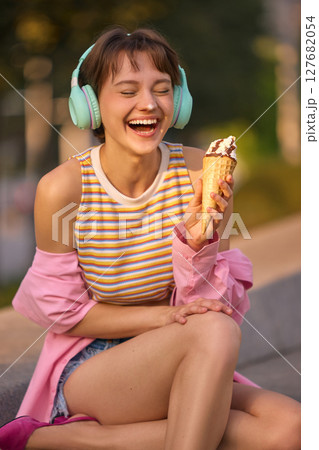 Sunshine bite. Woman enjoying ice cream and music outdoors in summer attire Sunshine bite. Woman enjoying ice cream and music outdoors in summer attire 127682054