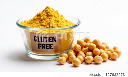 A labeled glass cup holds a mound of yellow chickpea flour marked gluten free, accompanied by raw chickpeas placed beside it on a clean white surface. 127682078