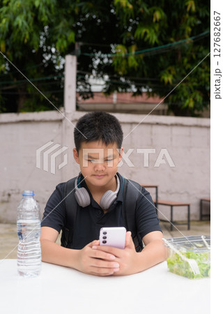 Thai boy eating salad and using wireless technology at outdoor table Thai boy eating salad and using wireless technology at outdoor table 127682667