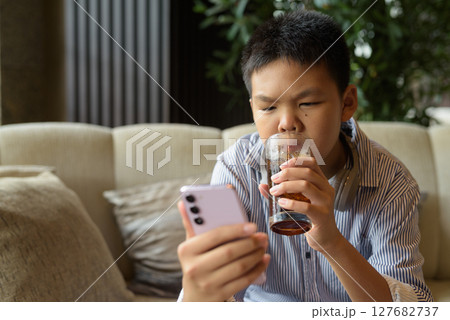 Thai boy drinking soft drink and using phone at cafe with headphones 127682737