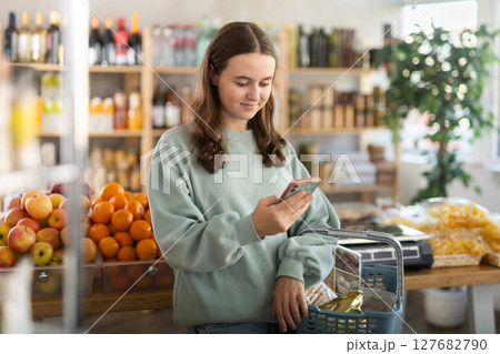 Visiting store, teen girl with phone is looking at something on shelf of showcase. Visiting store, teen girl with phone is looking at something on shelf of showcase. 127682790
