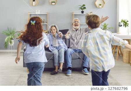 Grandchildren running to their senior happy smiling grandparent sitting on sofa at home to hug. 127682796
