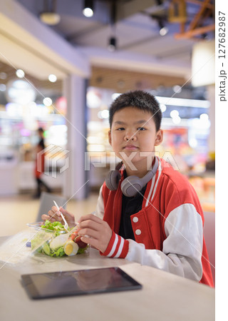 Teen Thai boy wearing headphones eating salad in cafeteria 127682987