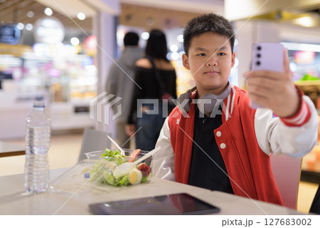 Teen Thai boy wearing headphones eating salad in cafeteria taking selfie with phone 127683002