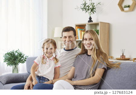 Cheerful family sitting on sofa with parents holding daughter on knees, making joyful home photo. Cheerful family sitting on sofa with parents holding daughter on knees, making joyful home photo. 127683124
