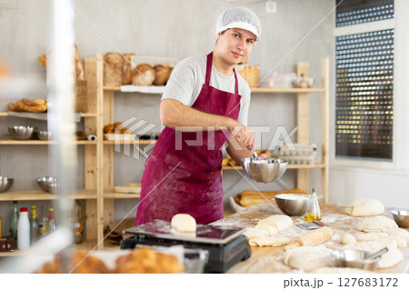 Guy worker in kitchen is stirring dough in bowl with whisk. 127683172