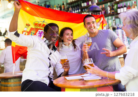 Happy multiracial sport fans holding and wrapping in a flag of Spain and supporting national football team in the bar 127683218
