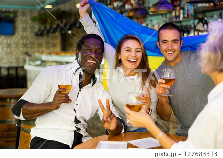 Happy friends of different ages drinking beer and celebrating the victory of the Ukrainian team in night bar Happy friends of different ages drinking beer and celebrating the victory of the Ukrainian team in night bar 127683313