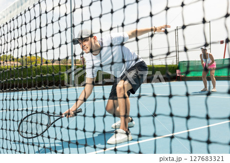 Portrait of man playing with male partner tennis doubles game on outdoors court. View through tennis net 127683321