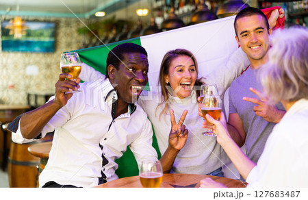 Excited diverse soccer supporters with flag of Italy celebrating victory with pint of beer in the pub 127683487