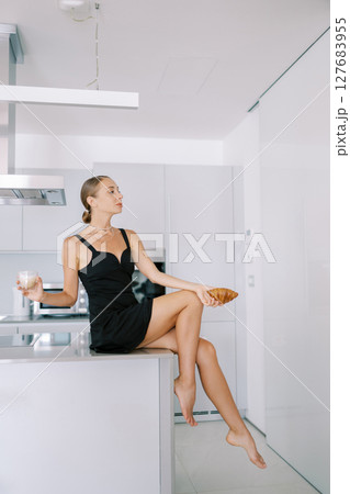 Young woman with a croissant and coffee in her hands sits on a table in the kitchen and looks away Young woman with a croissant and coffee in her hands sits on a table in the kitchen and looks away 127683955