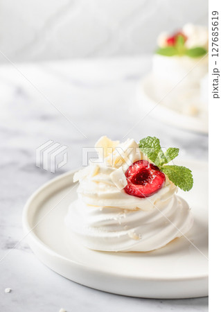 Meringues mini pavlova cake with whipped cream, fresh cherry and mint leaves on a plate, white marble background. Light summer dessert, text space Meringues mini pavlova cake with whipped cream, fresh cherry and mint leaves on a plate, white marble background. Light summer dessert, text space 127685619