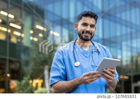 A friendly healthcare worker in blue scrubs holds a tablet while smiling, standing in front of a modern building, possibly a medical facility. 127685831