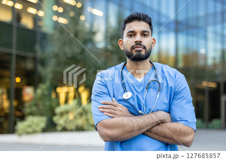 A healthcare professional stands outside, arms crossed, and is wearing scrubs and a stethoscope around his neck. The medical worker appears confident. 127685857