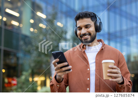 An attractive man listens to music while scrolling on his phone and holding a coffee cup. The background shows a modern building. An attractive man listens to music while scrolling on his phone and holding a coffee cup. The background shows a modern building. 127685874