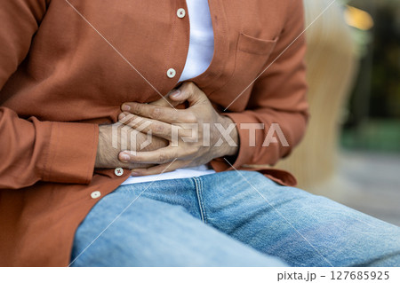 A man clutches his abdomen, suggesting abdominal discomfort or pain. The image highlights the pose, conveying a sense of physical distress or health concern. 127685925